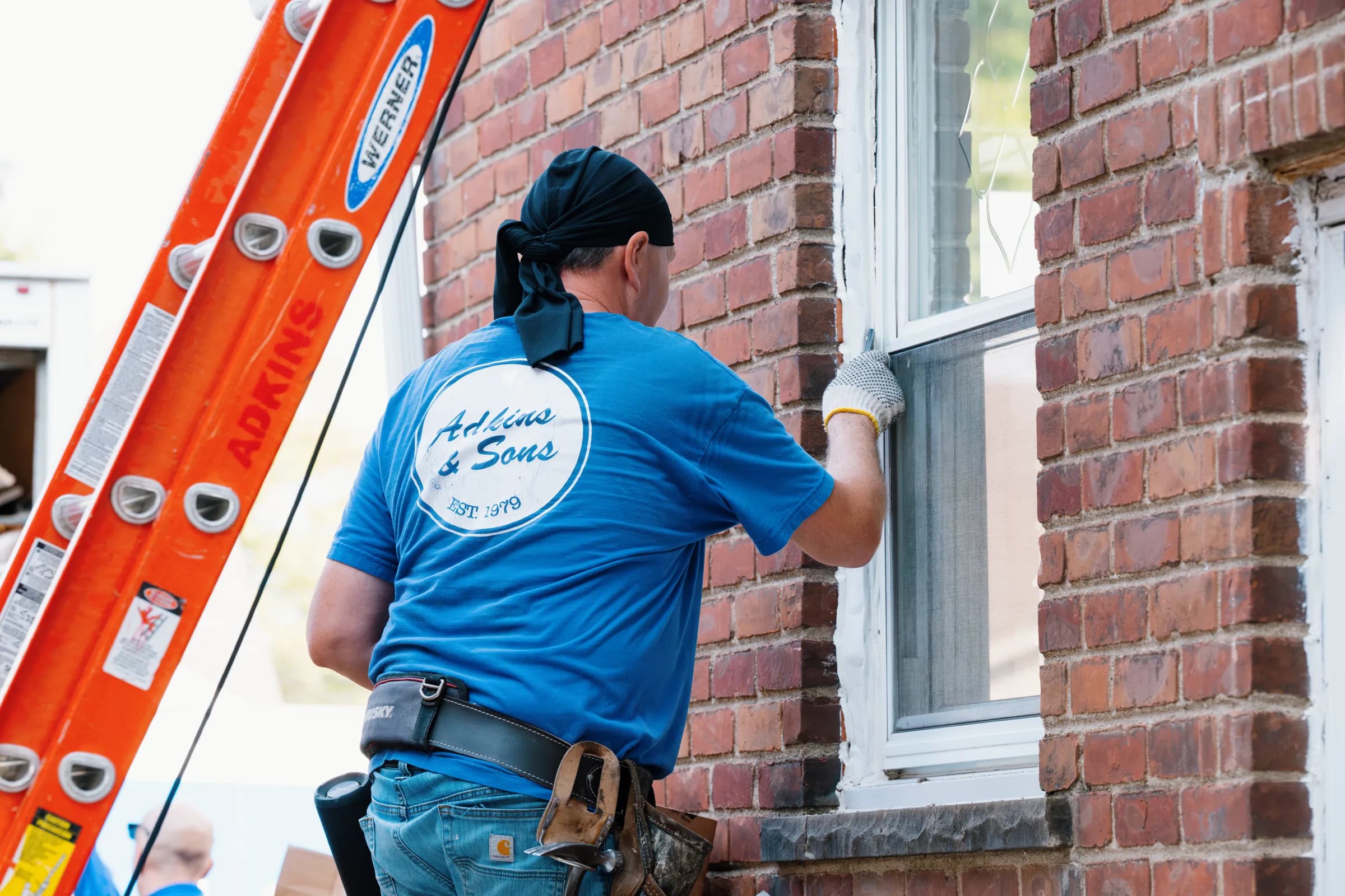An Adkins & Sons installer in a company shirt working a window opening from the company ladder
